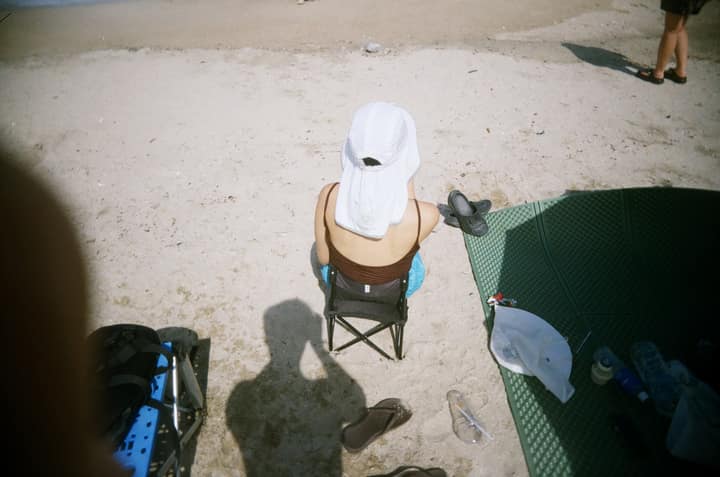 A person in a white sun hat seated on a beach, with the photographer's shadow in the sand